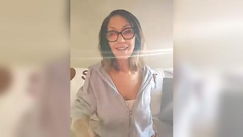 Media: Video of a smiling Asian woman with shoulder-length hair, wearing black-rimmed glasses and a light grey hoodie, standing in a dimly lit room.