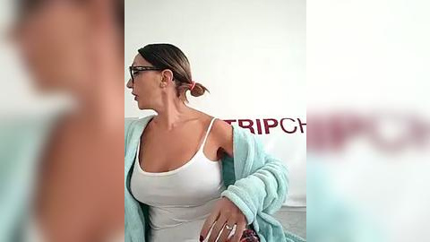 Media: Video of a woman in a white tank top and light blue robe, with glasses, dark hair tied back, standing indoors.