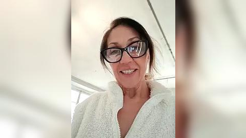 Media: Video of an elderly Caucasian woman with fair skin, short brown hair, and glasses, smiling in a white bathrobe. Background shows a bright, modern bathroom with a window.