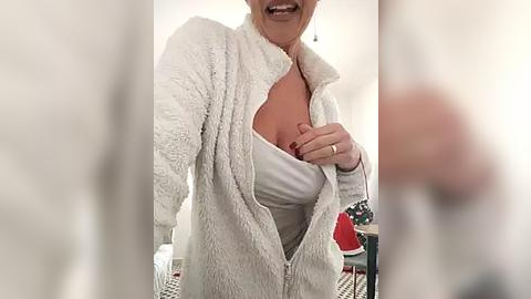 Media: Video of a middle-aged woman with fair skin, wearing a white, fuzzy robe, revealing a white bra underneath, in a brightly lit, modern room with white walls and a patterned floor.