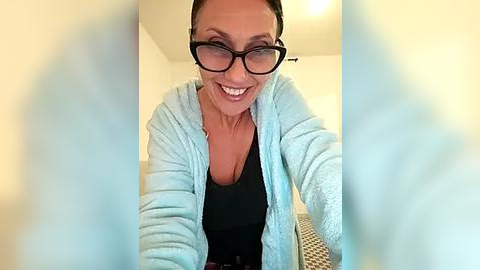 Media: Video of a smiling, light-skinned woman with glasses, medium-sized breasts, wearing a black top and light blue cardigan, taken from a low angle, with blurred foreground.