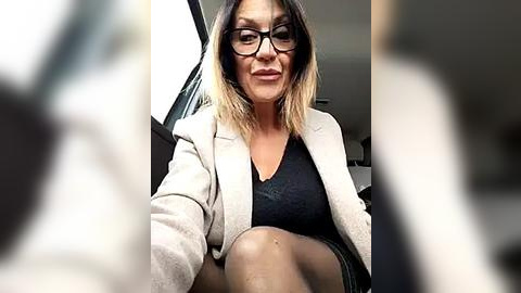 Media: Video of a woman with straightened, shoulder-length dark blonde hair, wearing black-rimmed glasses, a beige blazer over a black top, seated in a car. The background is blurred.