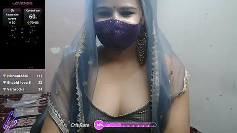 Media: A video of a light-skinned woman with medium-sized breasts, wearing a purple face mask and blue saree, standing against a plain wall. A live stream display in the background shows her username, views, and hearts.