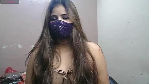 Media: Video of a young woman with long, straight brown hair, wearing a purple face mask and a beige lace bra. The background features a beige wall with peeling paint and a red object in the corner.