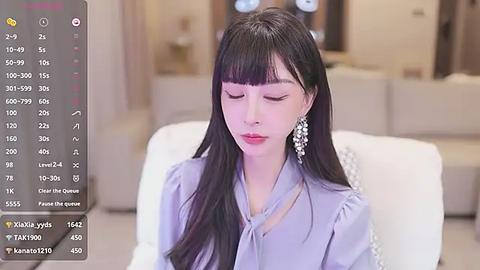 Media: Video of a young Asian woman with long black hair, wearing a light blue blouse, sitting in a modern living room.