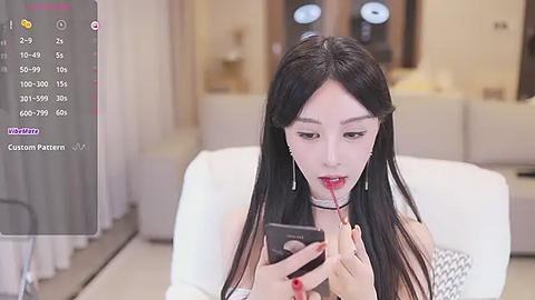 Media: Video of a young Asian woman with long black hair, fair skin, and red lipstick, applying lipstick while looking at a smartphone in a modern, white-lit living room.