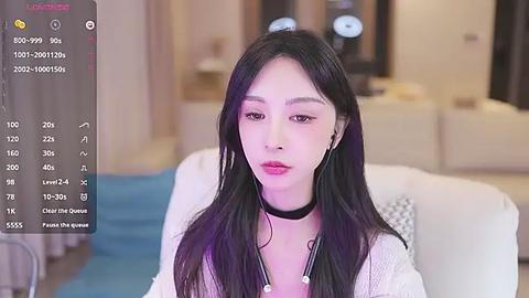 Media: A video of a young East Asian woman with long black hair, wearing a white knit cardigan and black choker, sitting on a white couch. The background is a modern, well-lit living room with soft furnishings.