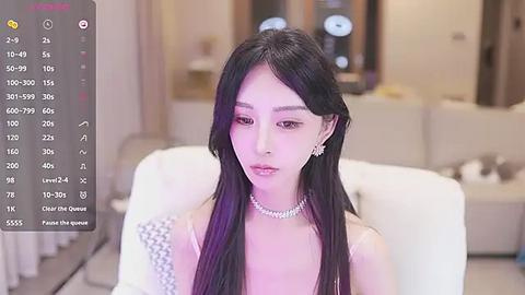 Media: Video of an East Asian woman with long black hair and fair skin, wearing a pearl necklace and a white strapless dress, seated in a modern, softly lit living room.