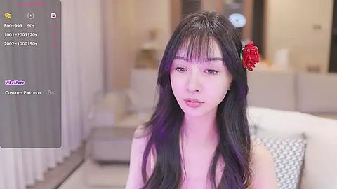 Media: Video of an Asian woman with long black hair, wearing a red flower, in a modern living room with neutral tones, featuring a gray sofa and beige curtains.