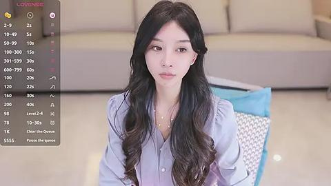 Media: Video of an East Asian woman with long, wavy black hair, wearing a light blue blouse, seated indoors. Background shows a beige sofa and a blue cushion.