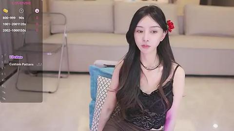 Media: Video of an East Asian woman with long black hair and a red flower in her hair, wearing a black lace dress, sitting on a blue chair in a modern living room.