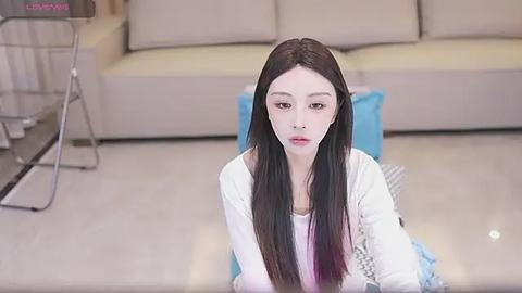 Media: Video of a young Asian woman with long black hair and pale skin, kneeling on a beige floor in a modern living room with light-colored furniture.