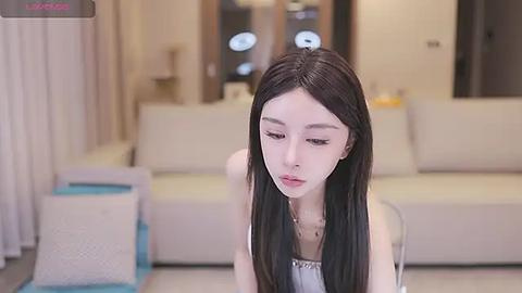 Media: Video of a young East Asian woman with long, straight black hair and fair skin, wearing a white top, seated in a modern, minimalist living room with beige furniture.