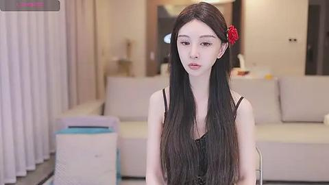 Media: Video of an East Asian woman with long black hair, fair skin, and red flower accessory, wearing a black sleeveless top, standing in a modern, minimalist living room with beige furniture.