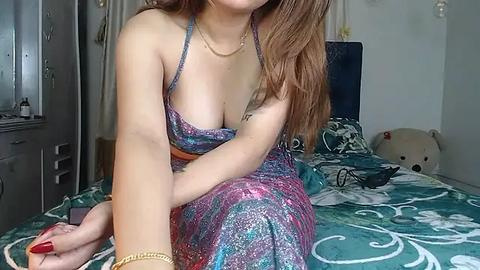 Media: Video of a light-skinned woman with long brown hair, wearing a low-cut, glittery dress, sitting on a bed with teal and white floral bedding. A stuffed animal and personal items are visible in the background.