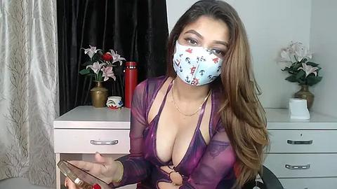 Media: Video of a light-skinned woman with long, wavy brown hair, wearing a blue face mask and a revealing purple lace bralette, sitting at a white dresser with flowers, a red candle, and a book.