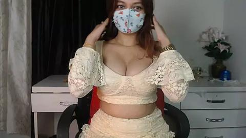 Media: Video of a young woman with medium skin tone, wearing a lace crop top and matching skirt, face mask, and holding her hair. Background features a white dresser, pink flowers, and dark curtains.