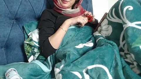 Media: Video of a woman lying on a teal blanket with white patterns, wearing a black top, red and white scarf, and light blue sneakers. She holds a smartphone, resting on a blue upholstered chair.