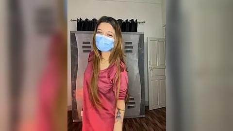 Media: Video of a young woman with long brown hair, wearing a pink t-shirt, blue surgical mask, and standing in front of metal lockers. Background includes a white door and dark curtains.