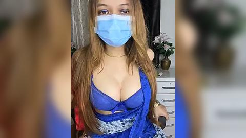 Media: Video of a woman in a blue face mask, revealing long brown hair, wearing a revealing blue lace bra, seated in a bedroom with a white dresser.