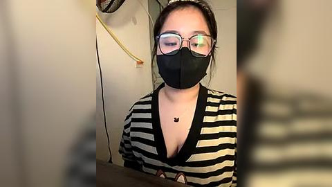 Media: Video of an East Asian woman with glasses, wearing a black mask and black-and-white striped shirt, standing in a bathroom with a visible sink.