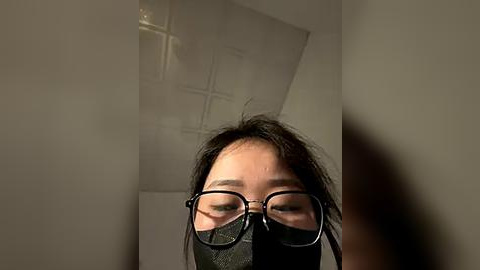 Media: A close-up video of a person with short black hair and glasses wearing a black face mask, standing in a dimly lit room with a glass block wall in the background.