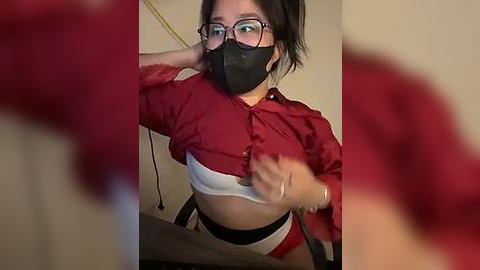 Media: Video of a woman with glasses and a black mask, lifting her red shirt to reveal a white bra, taken indoors with blurred background.