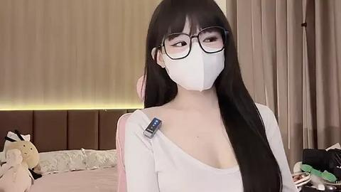 Media: Video of an Asian woman with long black hair, wearing glasses, a white mask, and a white off-shoulder top, sitting in a dimly lit bedroom with beige curtains and a bed.