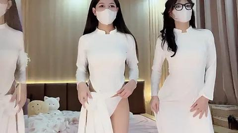 Media: Video of three East Asian women with long black hair, wearing white surgical masks and long-sleeved, high-necked gowns. One woman has glasses. Background shows a plush bed with teddy bears.