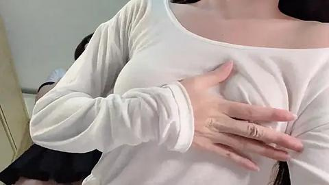 Media: Video of a light-skinned woman in a white long-sleeved top, left hand over her right breast, black skirt partially visible in the background.