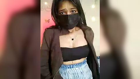 Media: Video of a young woman with long black hair, wearing a black face mask, black top, and light blue high-waisted shorts, standing indoors with blurred background.