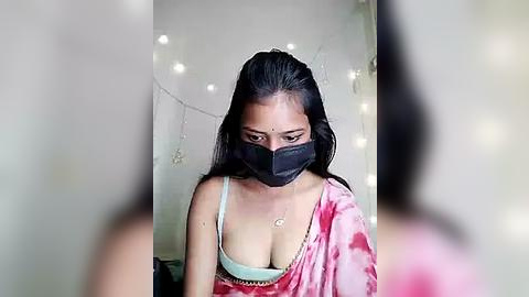 Media: Video of a South Asian woman with long black hair, wearing a pink floral-patterned sari, a black face mask, and a white bra. She has a light skin tone and is indoors with string lights in the background.