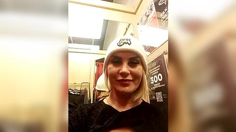 Media: A video of a blonde woman with light skin, wearing a black dress and a white beanie with a Star Wars logo, in a brightly lit, cluttered room with posters and a lamp.