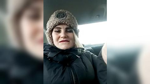 Media: A video of a smiling woman in a dark puffy coat and a beige knitted hat with a black logo, sitting in a car, with the background slightly blurred.