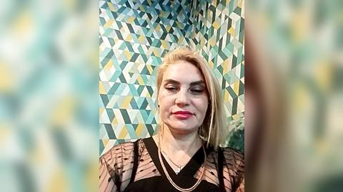 Media: Video of a smiling, fair-skinned, blonde woman with shoulder-length hair, wearing a sheer black top and gold necklace, standing against a geometric-patterned teal and yellow wall.