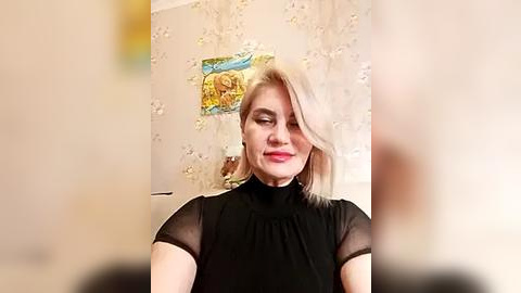 Media: Video of a smiling, light-skinned woman with short blonde hair, wearing a black sheer top, standing in front of a floral wallpaper with a yellow and green framed picture.