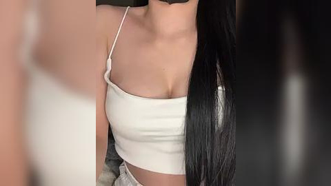 Media: Video of a young woman with long, straight black hair, wearing a white, off-shoulder crop top that accentuates her medium-sized breasts. She has fair skin and is seated against a blurred, dark background.