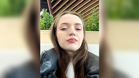 Media: A video of a young Caucasian woman with fair skin, straight brown hair, and light makeup. She wears a black leather jacket over a white top. Background features greenery and wooden slats.