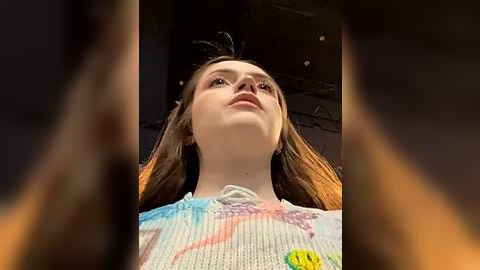 Media: Video of a young Caucasian woman with long, straight auburn hair, wearing a tie-dye sweater, looking upwards with a neutral expression. Background is blurred, possibly an indoor setting.