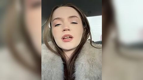 Media: A close-up video of a young woman with fair skin, brown hair, and full lips, wearing a fur coat, taken from a slightly blurred, out-of-focus perspective.