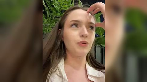 Media: A video of a young Caucasian woman with long brown hair, wearing a beige blouse, having her forehead touched by another hand. Background features lush green leaves, creating a serene, natural ambiance.