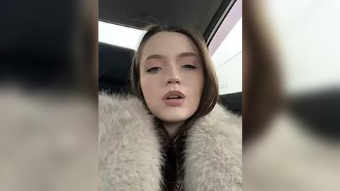 Media: A video of a young woman with fair skin, light brown hair, and pink makeup, wearing a fluffy white fur coat, seated in a car with blurred faces in the foreground.