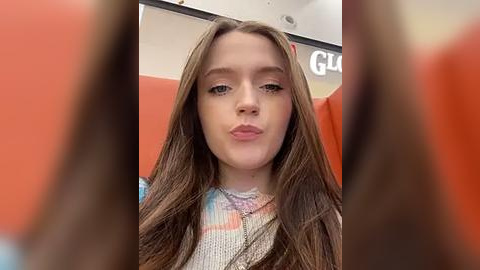 Media: A video of a young Caucasian girl with long brown hair, wearing a colorful, patterned sweater, pouting playfully in an orange-themed room with blurred figures in the background.