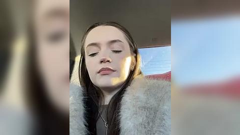 Media: A video of a young woman with fair skin and long, dark hair, wearing a gray fur coat, seated in a car with sunlight streaming in.