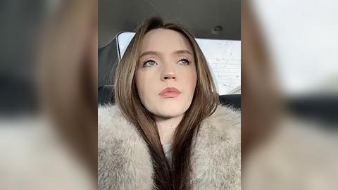 Media: Video of a young woman with fair skin, long brown hair, and green eyes, wearing a white coat, seated in a car with blurred background.