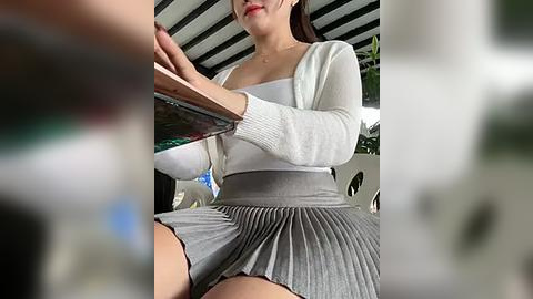 Media: A video of an Asian woman with fair skin, wearing a white cardigan over a white top and pleated gray skirt, sitting indoors with a blurred background.