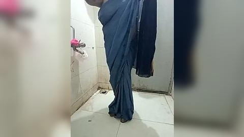 Media: Video of a person, presumably female, wearing a dark blue sari, standing in a small, tiled bathroom with a pink towel hanging on the wall and a dark garment draped over a towel rack.