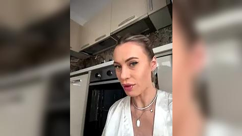 Media: Video of a Caucasian woman with fair skin, light brown hair, and green eyes, wearing a white satin robe, standing in a modern kitchen with beige cabinets and a stainless steel oven.