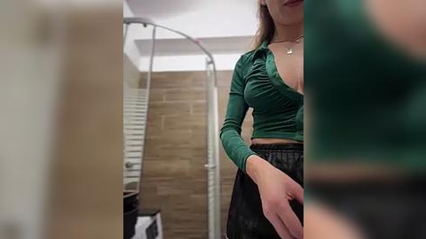 Media: Video of a woman in a green, low-cut, long-sleeved top and high-waisted black skirt, standing in a modern bathroom with a beige tiled wall and shower curtain.