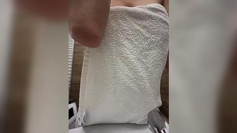 Media: Video of a person's hand holding a white, textured bath towel wrapped around their waist. Background features a beige tiled bathroom wall and partially visible white blinds.